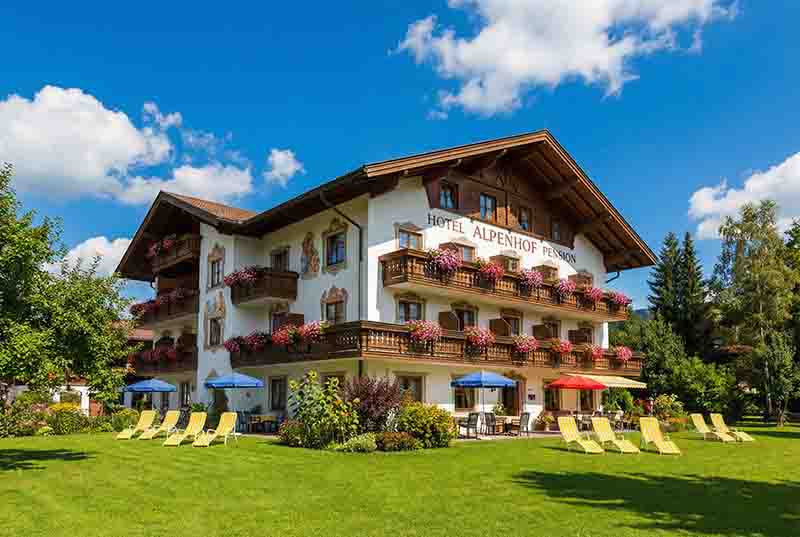 Traditionelles Hotel Alpenhof Pension im bayerischen Stil mit weißer Fassade, kunstvollen Wandmalereien und blumengeschmückten Holzbalkonen. 