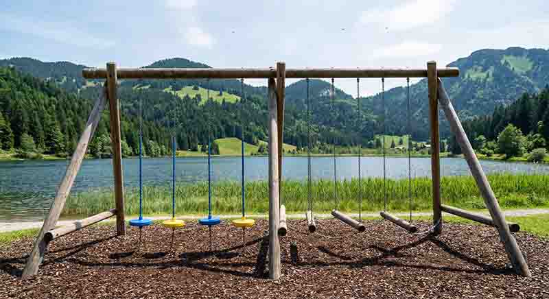 Ein Abenteuerspielplatz aus Holz vor der Kulisse eines idyllischen Oberbayrischen Bergsees und bewaldeten Alpengipfeln unter einem hellen Sommerhimmel.