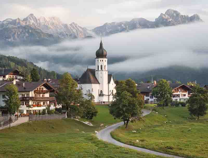 Ein idyllisches Dorf in Garmisch-Partenkirchen vor einer Kulisse aus hohen, teilweise wolkenverhangenen Bergen. Im Zentrum steht eine weiße Kirche mit einer markanten dunklen Zwiebelhaube, umgeben von grünen Wiesen und vereinzelten Bäumen im Vordergrund.