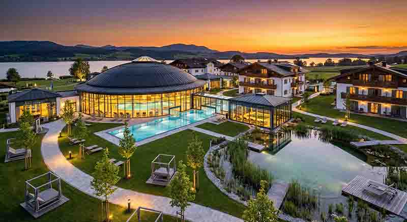 Eine weitläufige, luxuriöse Wellness-Resort-Anlage in Oberbayern bei stimmungsvoller Abenddämmerung an einem See.