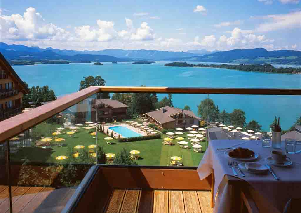 Blick von einem Hotelbalkon in Oberbayern mit gedecktem Frühstückstisch auf eine Hotelanlage mit Pool, grüne Liegewiesen mit gelben Sonnenschirmen und den Chiemsee vor bayerischer Alpenkulisse.