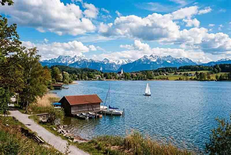 Starnberger See, Fuenfseenland, bei Sonnenschein. Ein hölzernes Bootshaus mit mehreren kleinen Booten liegt am Steg, während ein Segelboot auf dem Wasser kreuzt. Im Hintergrund erstreckt sich eine teils schneebedeckte Gebirgskette