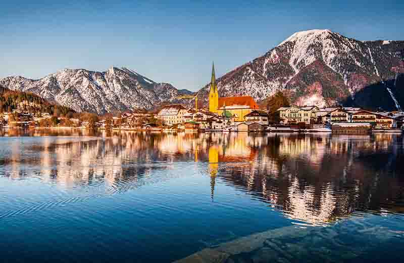 Rottach-Egern am Tegernsee. Im Vordergrund spiegelt sich das klare, blaue Wasser des Sees, während im Hintergrund die markante Pfarrkirche St. Laurentius und die bayerischen Alpen unter einem strahlend blauen Himmel zu sehen sind.